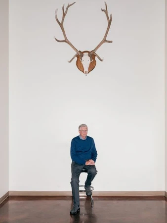 Elderly man in blue sweater seated beneath deer antlers on a stool in a minimalist interior, portrait-style, high-key lighting.