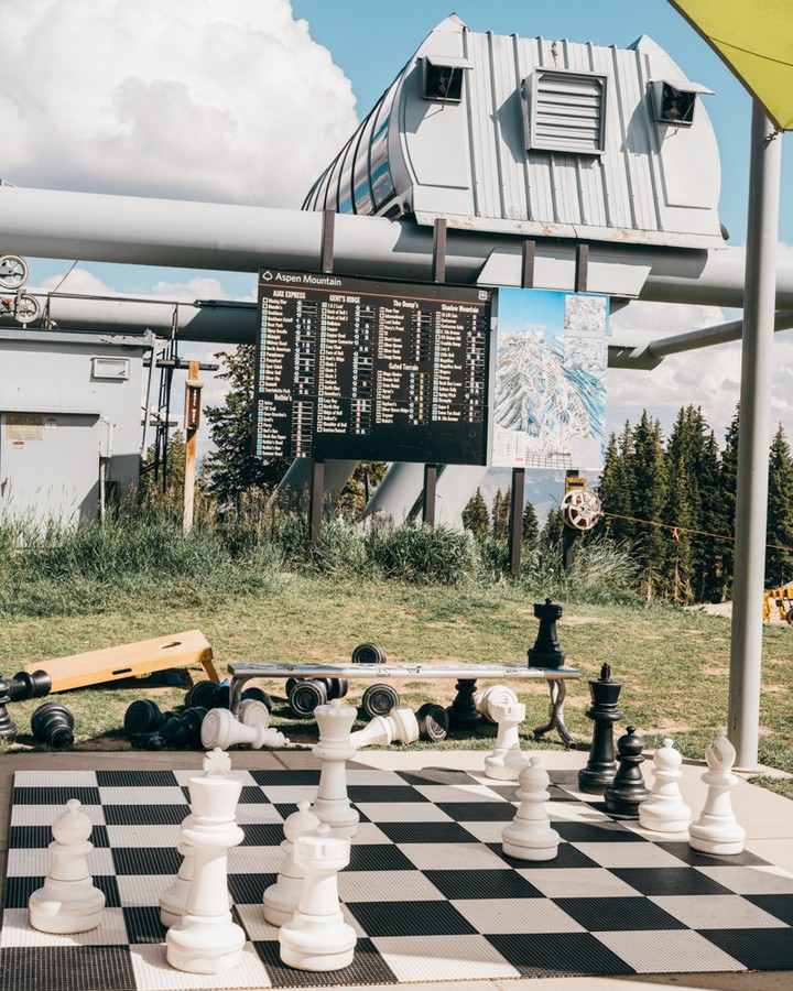 Outdoor chessboard with large pieces in a grassy park with ski lift infrastructure and mountain views on a sunny day at a ski resort.