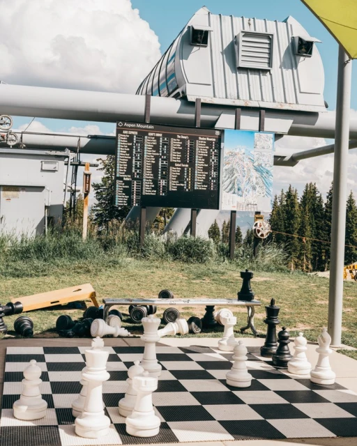 Outdoor chessboard with large pieces in a grassy park with ski lift infrastructure and mountain views on a sunny day at a ski resort.