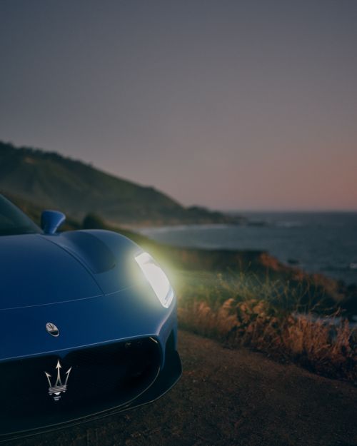 A blue Maserati sports car is parked on a coastal road with headlights on, showcasing its sleek design against the ocean backdrop.