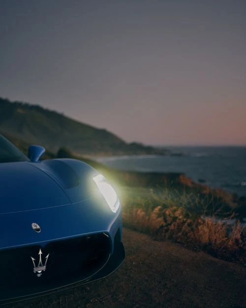 A blue Maserati sports car is parked on a coastal road with headlights on, showcasing its sleek design against the ocean backdrop.