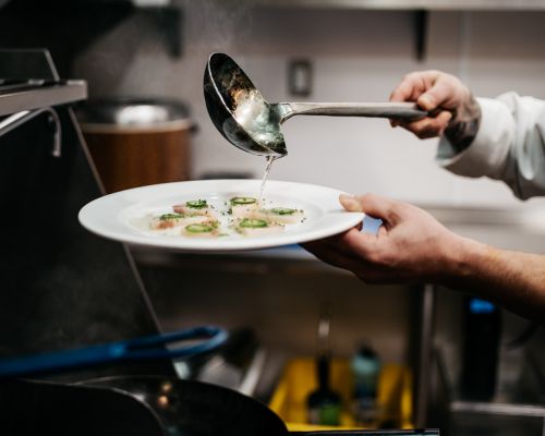 Chef pours sauce over scallop dish with jalapeños. Close-up captures professional food preparation in a commercial kitchen setting.