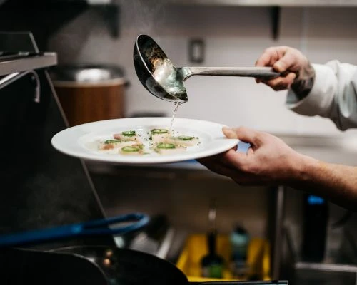 Chef pours sauce over scallop dish with jalapeños. Close-up captures professional food preparation in a commercial kitchen setting.