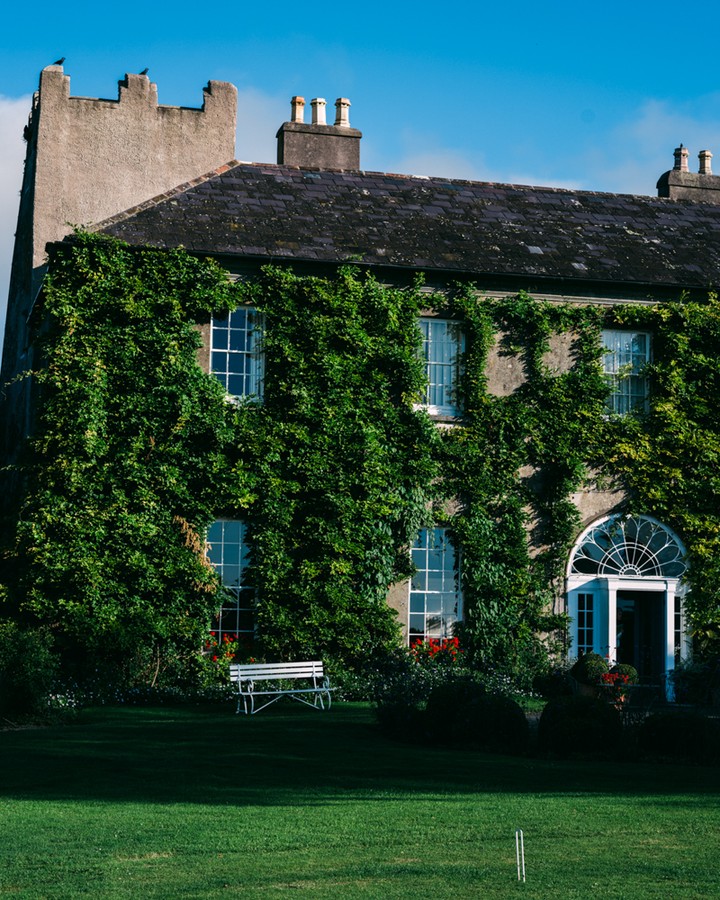 Exterior of a stone building covered in ivy, with white windows and doors, a bench on the lawn, and a blue sky above.