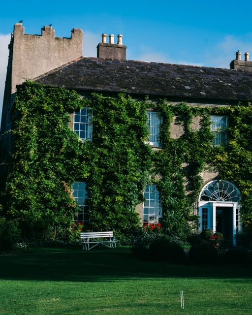 Exterior of a stone building covered in ivy, with white windows and doors, a bench on the lawn, and a blue sky above.