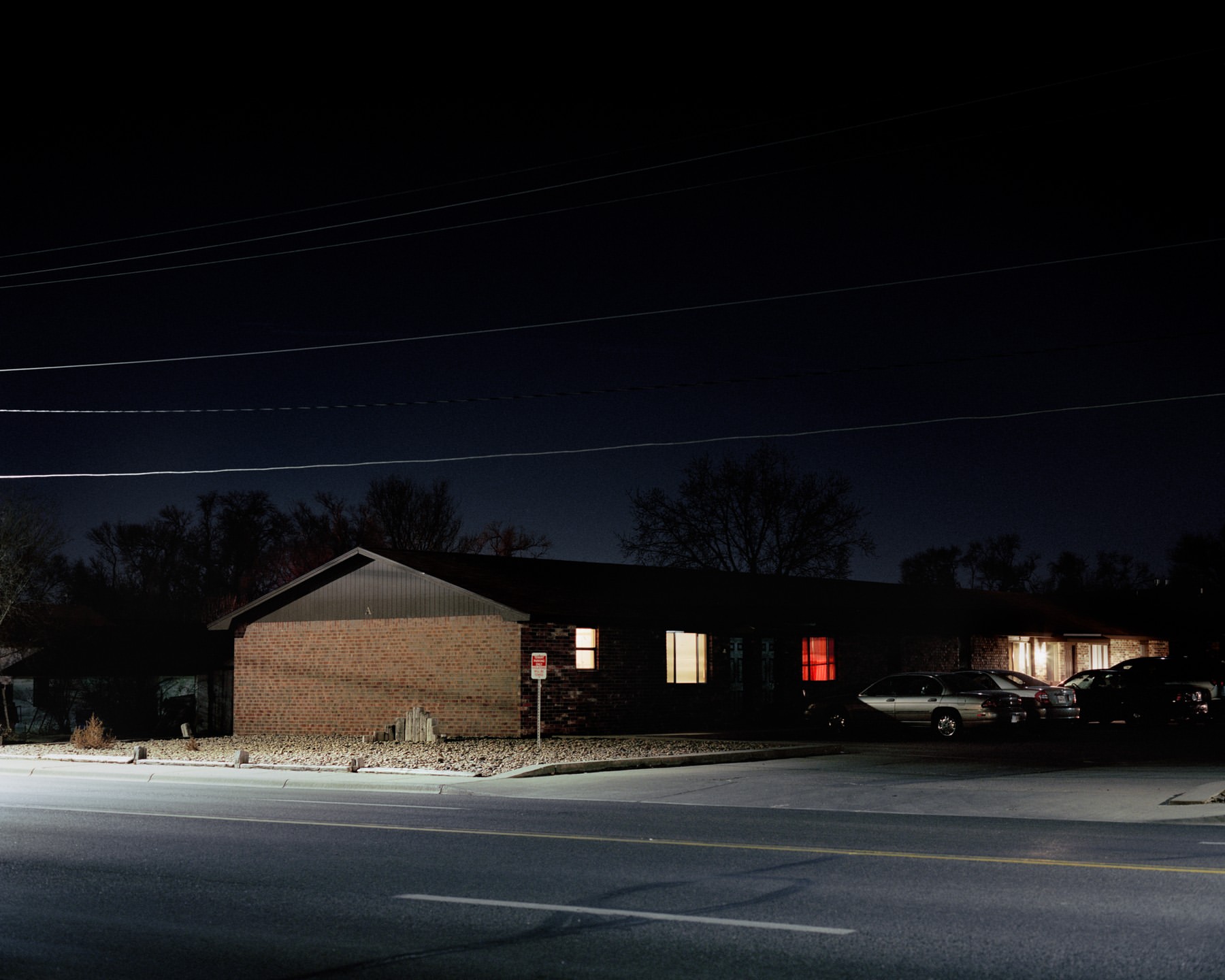 Night scene showcasing a one-story brick building with illuminated windows, accompanied by parked cars and a dark sky backdrop. 