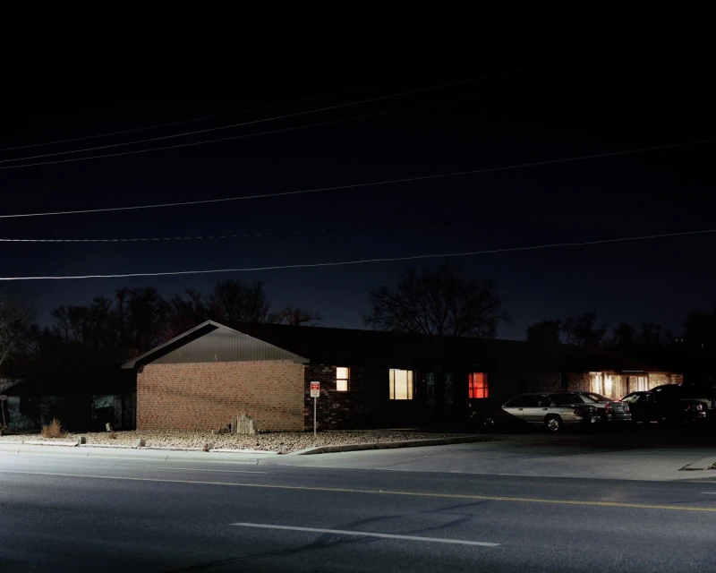 Night scene showcasing a one-story brick building with illuminated windows, accompanied by parked cars and a dark sky backdrop. 