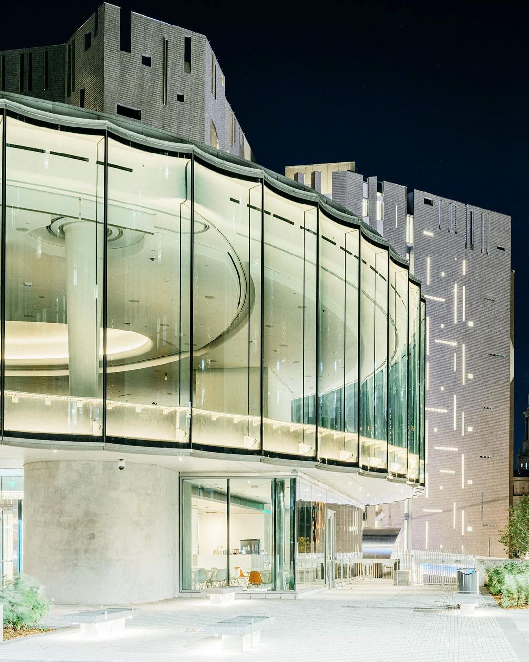 Exterior view of a modern building with glass walls and geometric concrete elements at night, reflecting interior lighting.