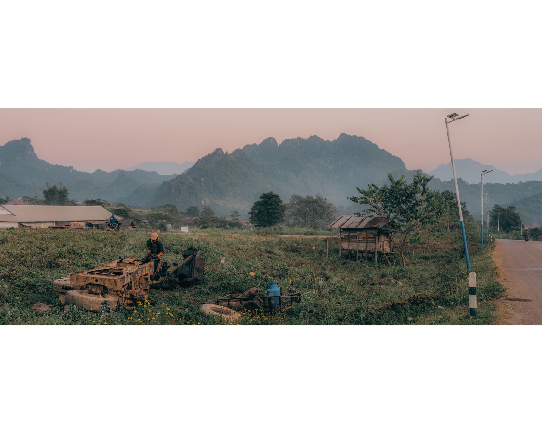 Rural landscape featuring a person atop aged machinery in a lush field, set against a backdrop of distant mountains and a rustic village.