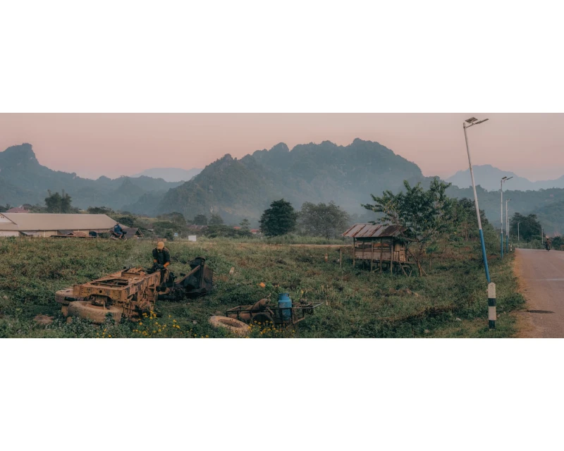 Rural landscape featuring a person atop aged machinery in a lush field, set against a backdrop of distant mountains and a rustic village.