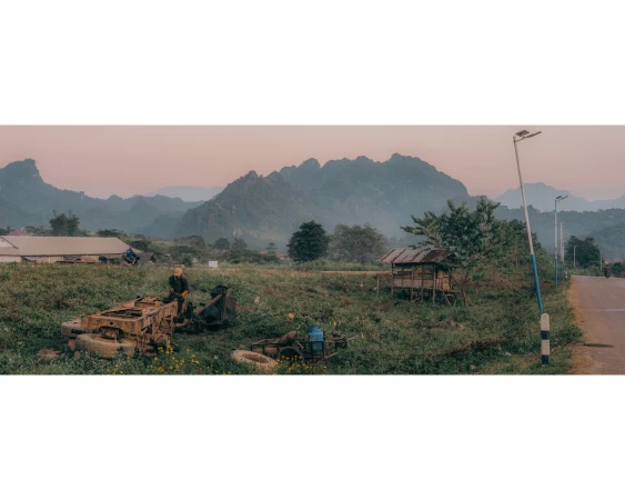 Rural landscape featuring a person atop aged machinery in a lush field, set against a backdrop of distant mountains and a rustic village.