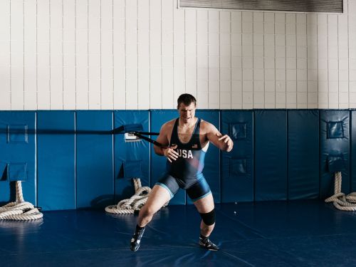 A male athlete in wrestling uniform with USA logo, training with resistance band in a sports facility with blue and white walls.