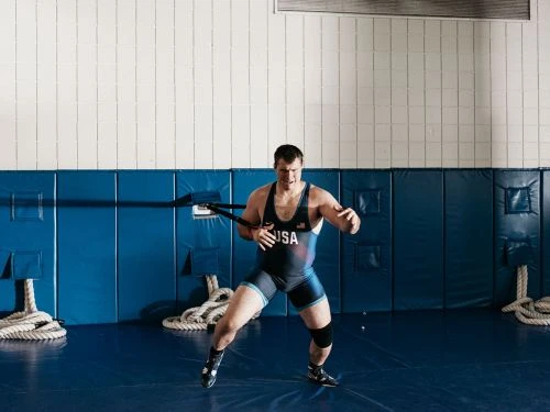 A male athlete in wrestling uniform with USA logo, training with resistance band in a sports facility with blue and white walls.