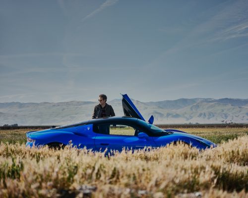 A blue Maserati sports car is parked in a field with a man standing beside it, under a blue sky with distant mountains.