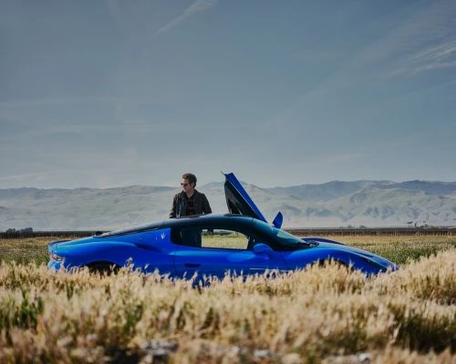 A blue Maserati sports car is parked in a field with a man standing beside it, under a blue sky with distant mountains.
