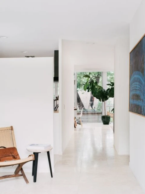 Interior shot of a hallway with white walls, a chair, a small table, a houseplant, and a blue painting in a minimalist style.