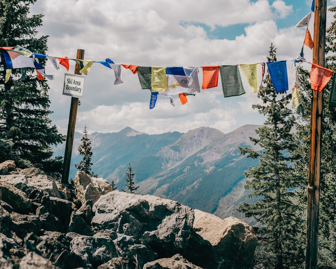 Mountain scenery with colorful prayer flags strung between trees, sign indicating a ski area boundary, and partly cloudy sky.