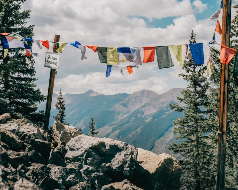 Mountain scenery with colorful prayer flags strung between trees, sign indicating a ski area boundary, and partly cloudy sky.