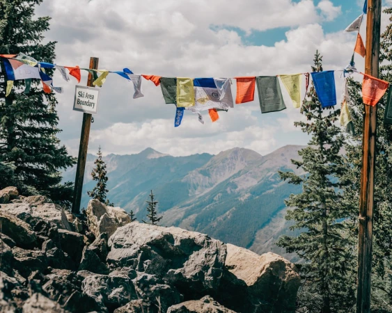 Mountain scenery with colorful prayer flags strung between trees, sign indicating a ski area boundary, and partly cloudy sky.