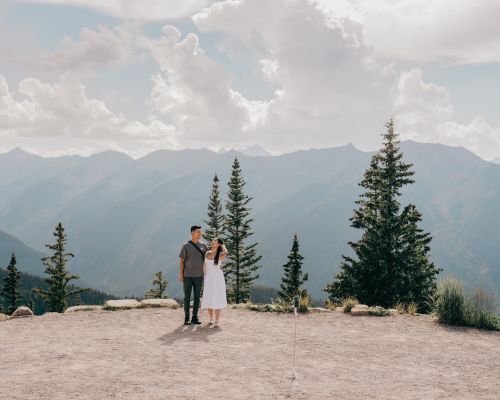 A couple stands together on a mountain overlook with a scenic view of mountain ranges and cloudy skies in the background.