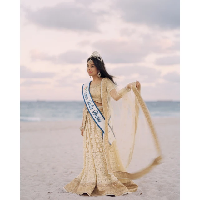 Young woman in traditional Indian dress stands on a sandy beach with the ocean and sky in the background, wearing a tiara and sash.