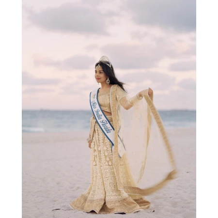 Young woman in traditional Indian dress stands on a sandy beach with the ocean and sky in the background, wearing a tiara and sash.