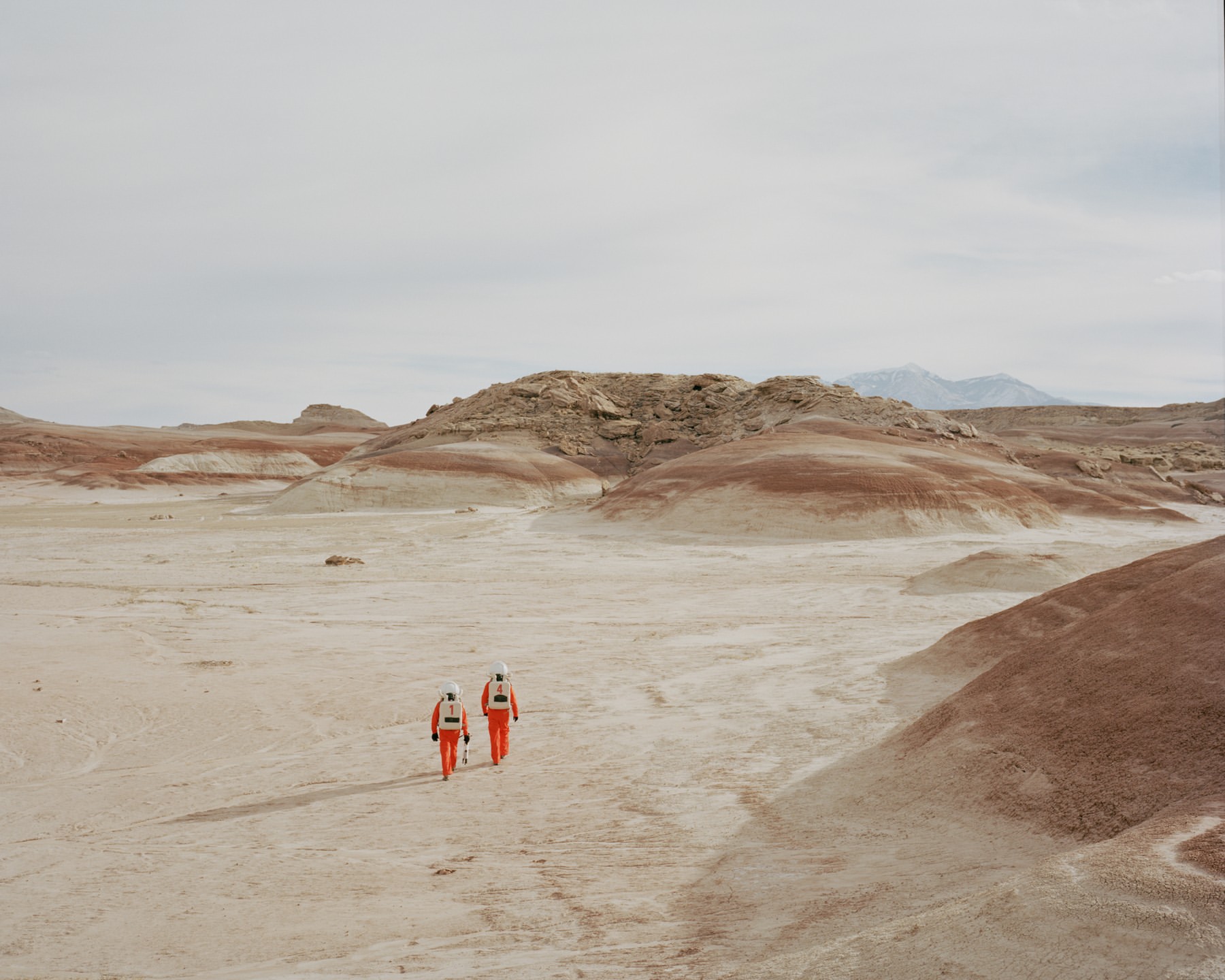 Two people in orange spacesuits walk across a barren, sandy landscape with layered rock formations under a cloudy sky, creating a scene reminiscent of Mars.