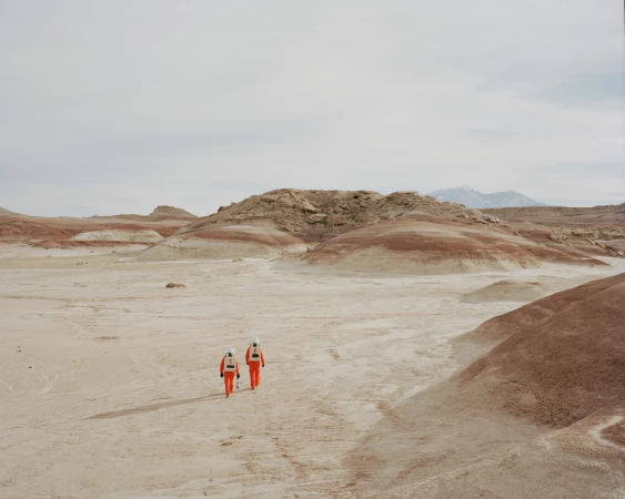Two people in orange spacesuits walk across a barren, sandy landscape with layered rock formations under a cloudy sky, creating a scene reminiscent of Mars.