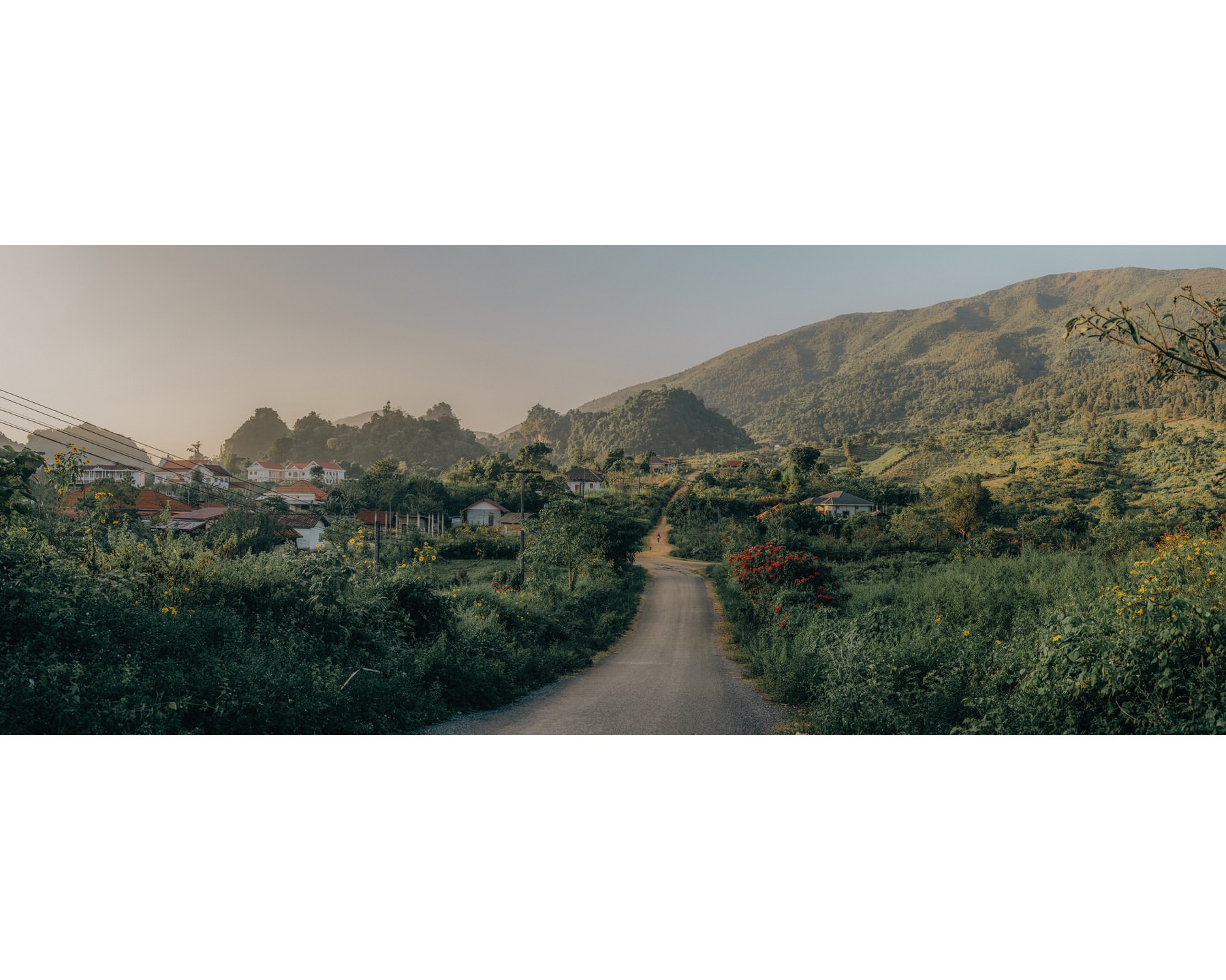 Scenic view of a rural road leading through lush greenery towards a village nestled between rolling hills and distant mountains.
