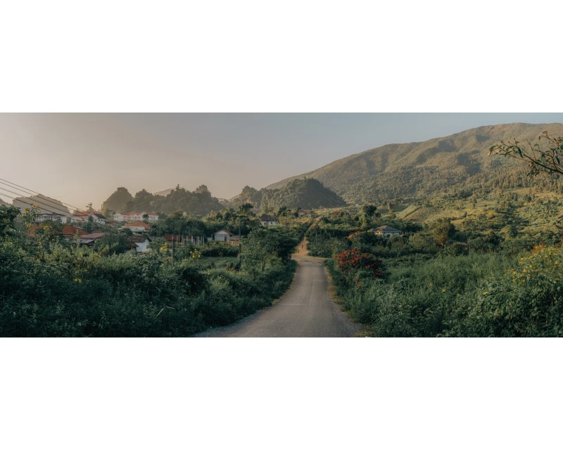 Scenic view of a rural road leading through lush greenery towards a village nestled between rolling hills and distant mountains.