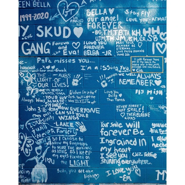 Brick wall mural with white handwritten text, love messages, names, and drawings in blue, white, and gold paint, memorial art.