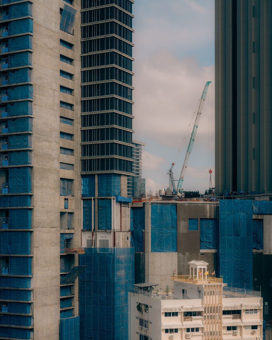 View of high-rise building construction site with a crane, concrete structures, and blue safety netting against a bright, cloudy sky.