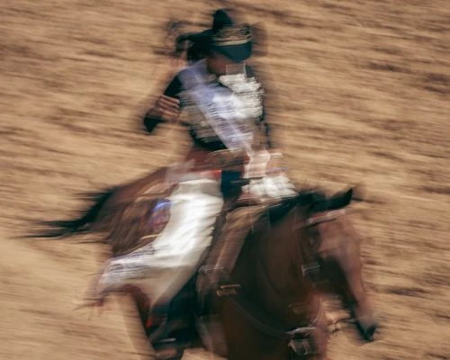 A female equestrian in traditional dress rides a horse at speed, captured with motion blur in an outdoor, sunlit arena filled with sand.