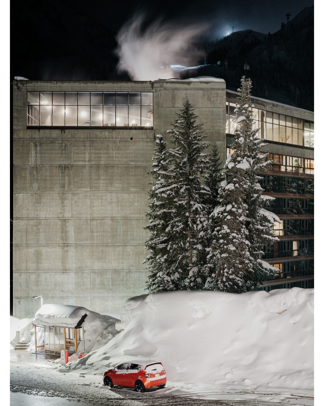 Exterior of a concrete building at night with snow-covered evergreen trees, a small shelter, and a red car parked nearby.