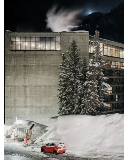 Exterior of a concrete building at night with snow-covered evergreen trees, a small shelter, and a red car parked nearby.