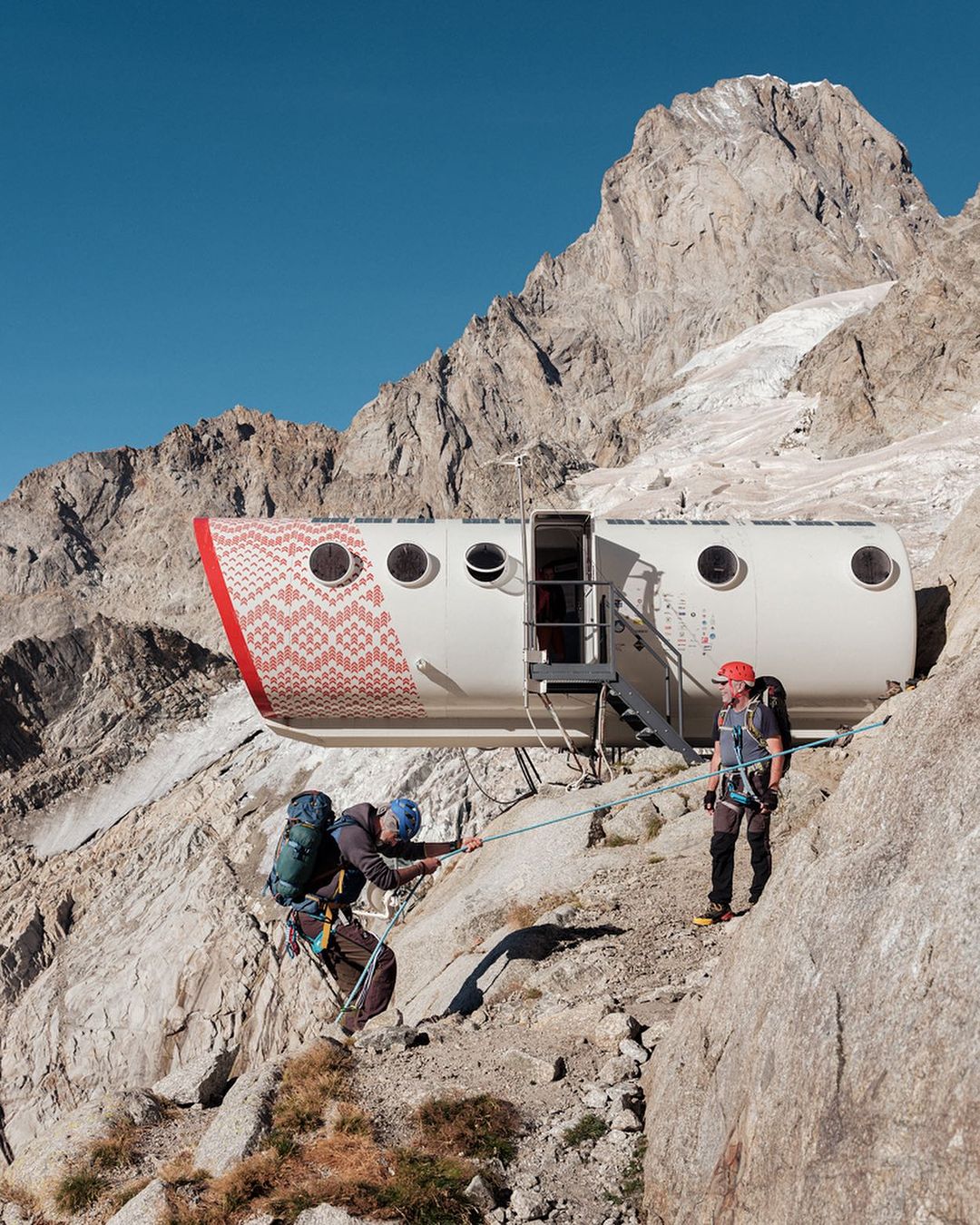 Two climbers ascend a rocky slope with a rope, near a mountain shelter in the mountains against a blue sky on a sunny day.