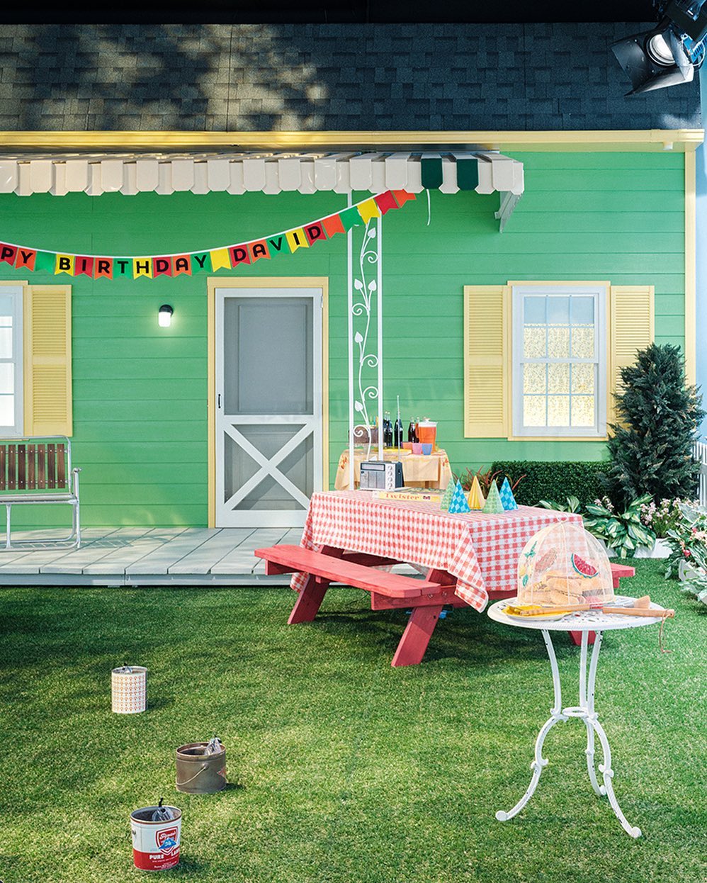 Outdoor birthday scene with a green house, red picnic table, party hats, and painted cans on the lawn on a sunny day.