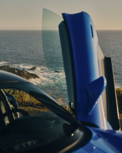 Blue car with a gull-wing door open, parked on a cliff overlooking the ocean. Bright sunlight, waves, and rocky coastline visible.