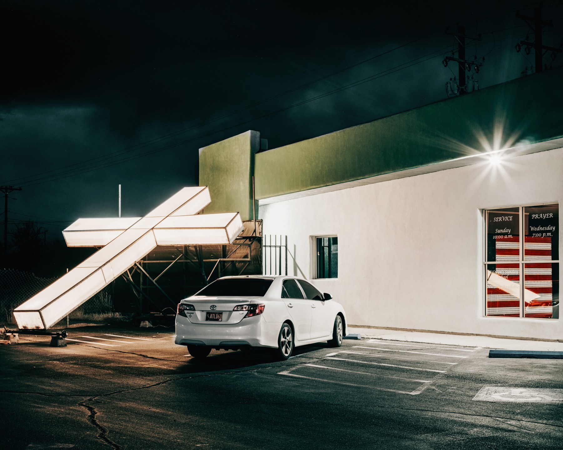 Nighttime parking lot scene with a white sedan parked next to an illuminated cross sign, near a building with window signage.