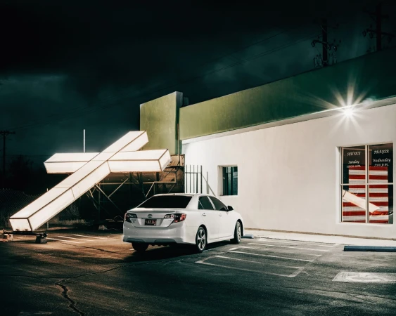 Nighttime parking lot scene with a white sedan parked next to an illuminated cross sign, near a building with window signage.