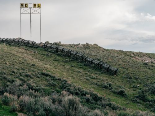 Rural scene featuring a grassy hillside and wooden fence leading to an old motel sign against an overcast sky.