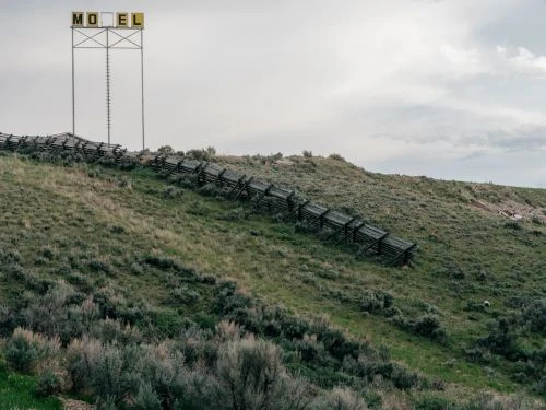 Rural scene featuring a grassy hillside and wooden fence leading to an old motel sign against an overcast sky.