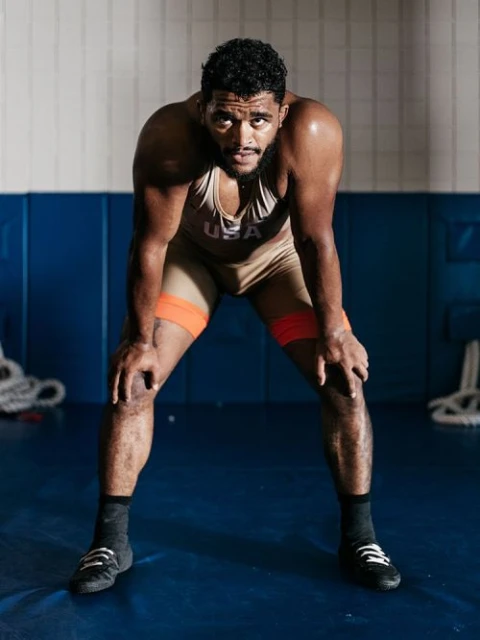 Wrestler in beige singlet with USA logo, resting with hands on knees on blue mat in wrestling gym with white wall and equipment.