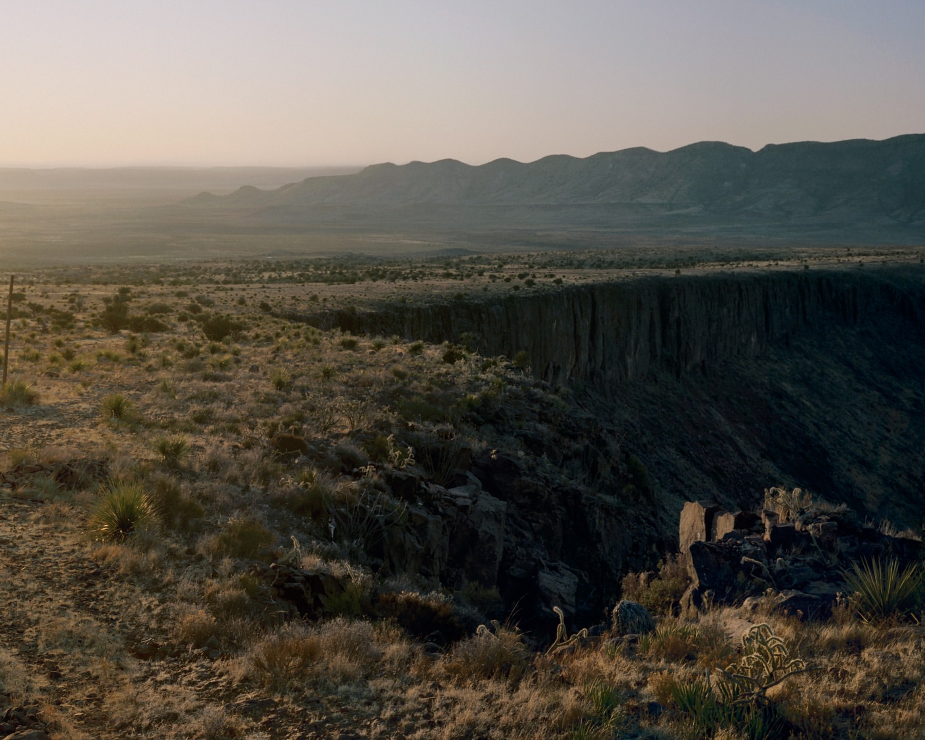 Expansive desert landscape view with a rugged canyon edge, arid vegetation, and distant mountains under a soft, hazy sky at dusk.