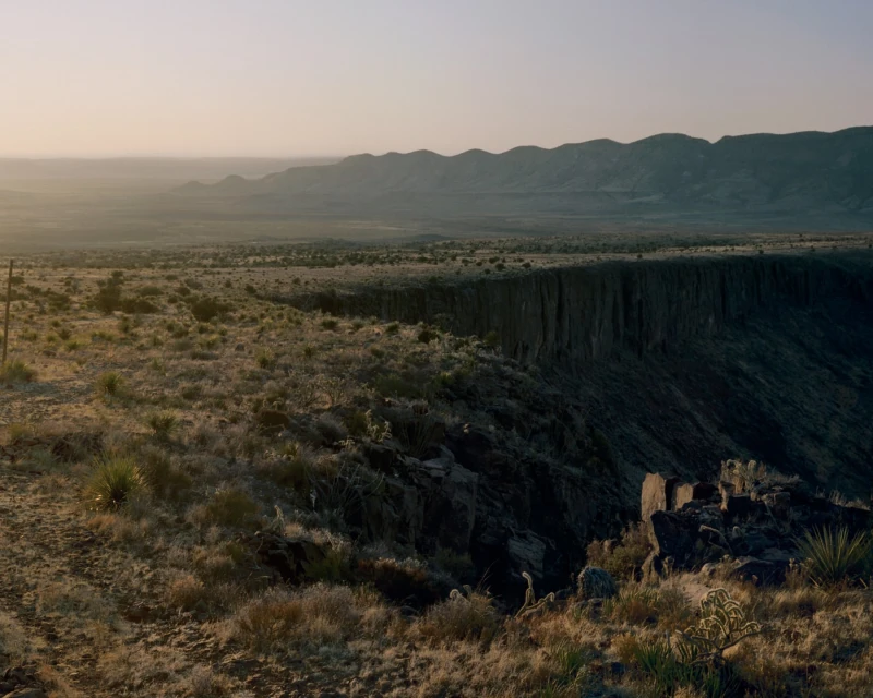 Expansive desert landscape view with a rugged canyon edge, arid vegetation, and distant mountains under a soft, hazy sky at dusk.