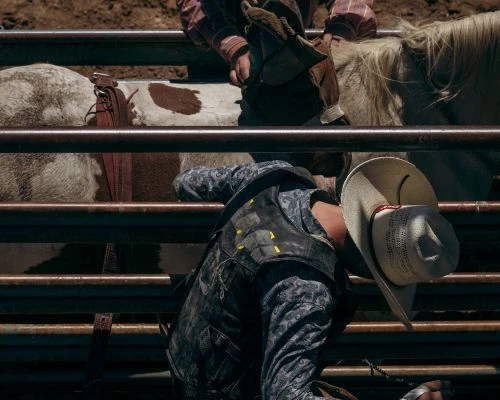 Cowboy riding a bull in a rodeo, wearing a cowboy hat and protective gear, captured in an outdoor arena with metal fencing.