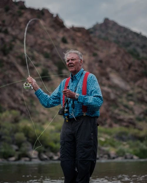 Elderly man fly-fishing in a river, wearing a blue plaid shirt and red suspenders, with a mountain backdrop and fly rod in hand.