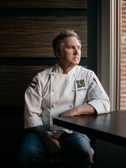 Portrait of a chef in a white uniform, sitting at a dark table looking out a window with brick wall, medium shot, indoors.