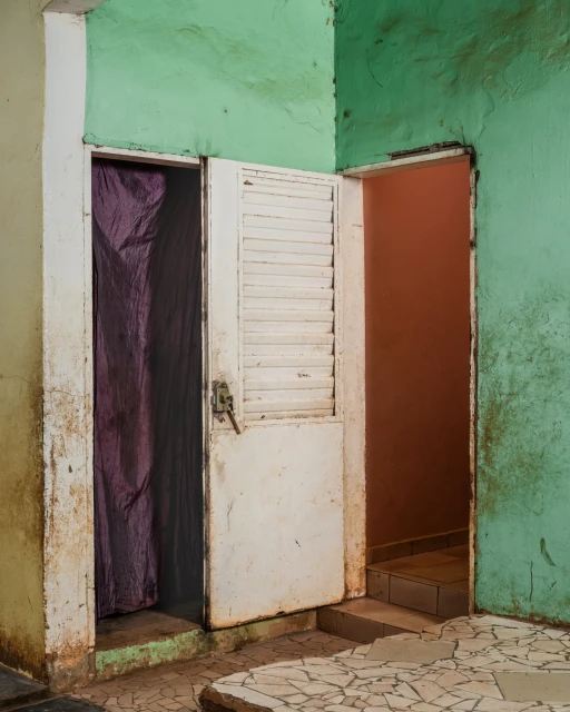 Interior view with a weathered white door ajar, purple curtain, and a glimpse into an adjacent room with tiled steps.