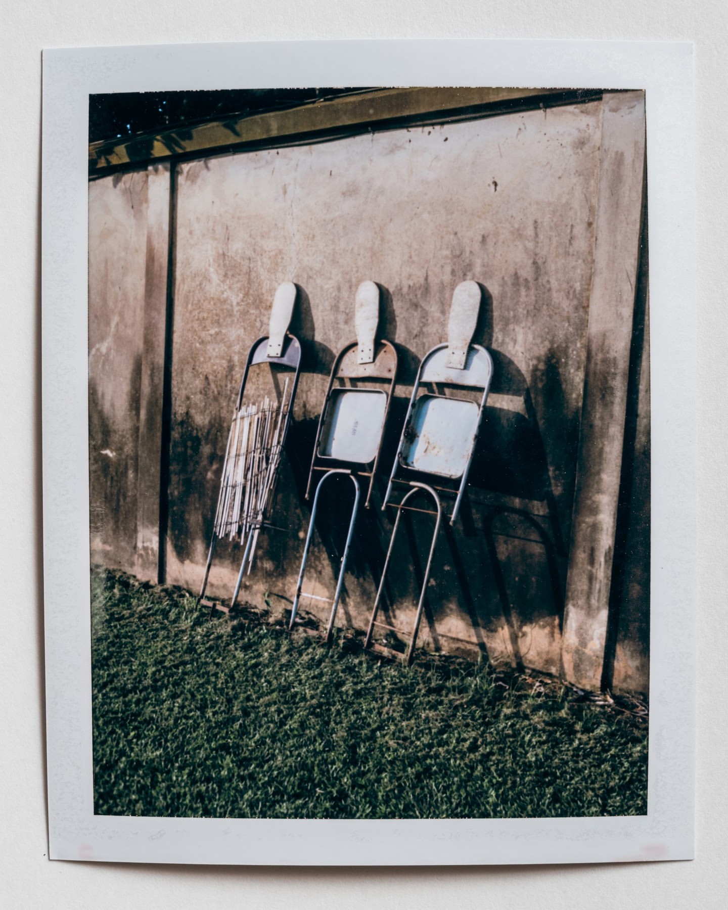 Three metal folding chairs stand against a concrete wall outdoors on a sunny day, casting shadows on the wall and green grass below.